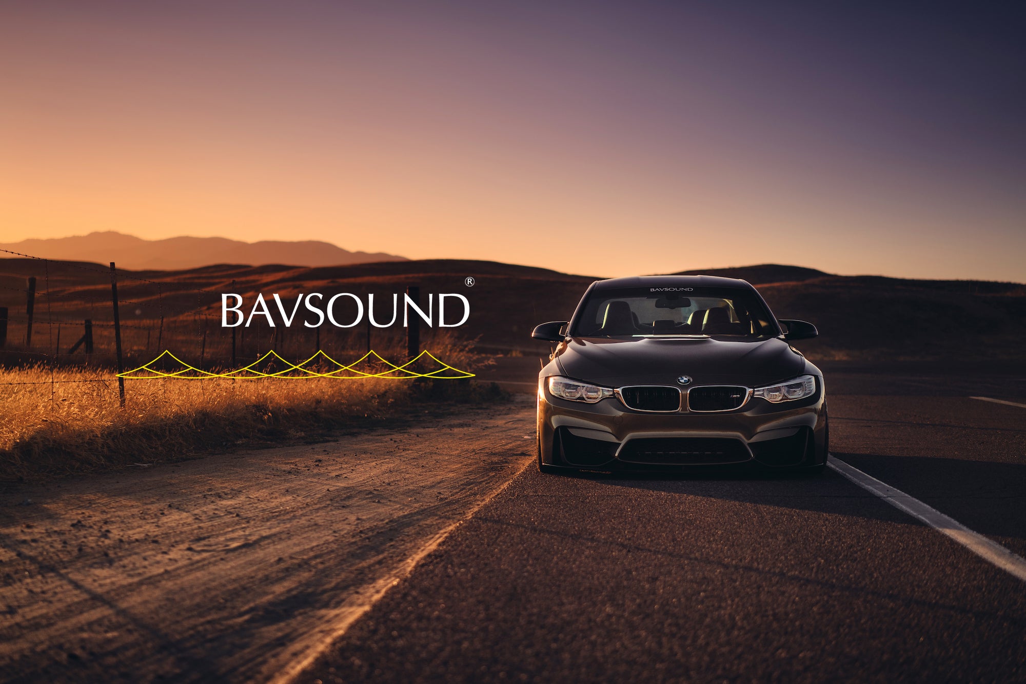 A Bronze Gray BMW sitting roadside at sunset next to the Bavsound logo. 